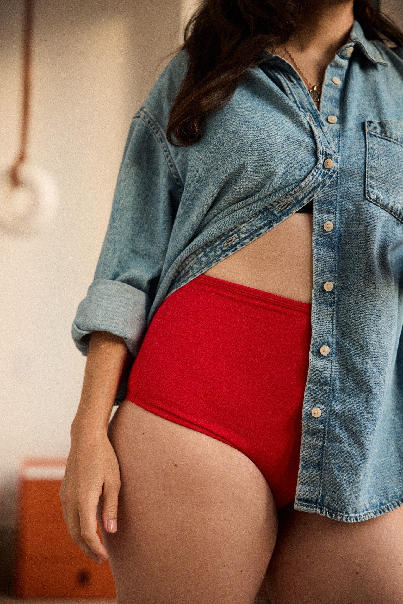 Woman in her 40s wearing a denim shirt over a pair of red underwear in a bedroom. The underwear is sweat wicking and made from bamboo to help with perimenopause symptoms. It also has leak protection.